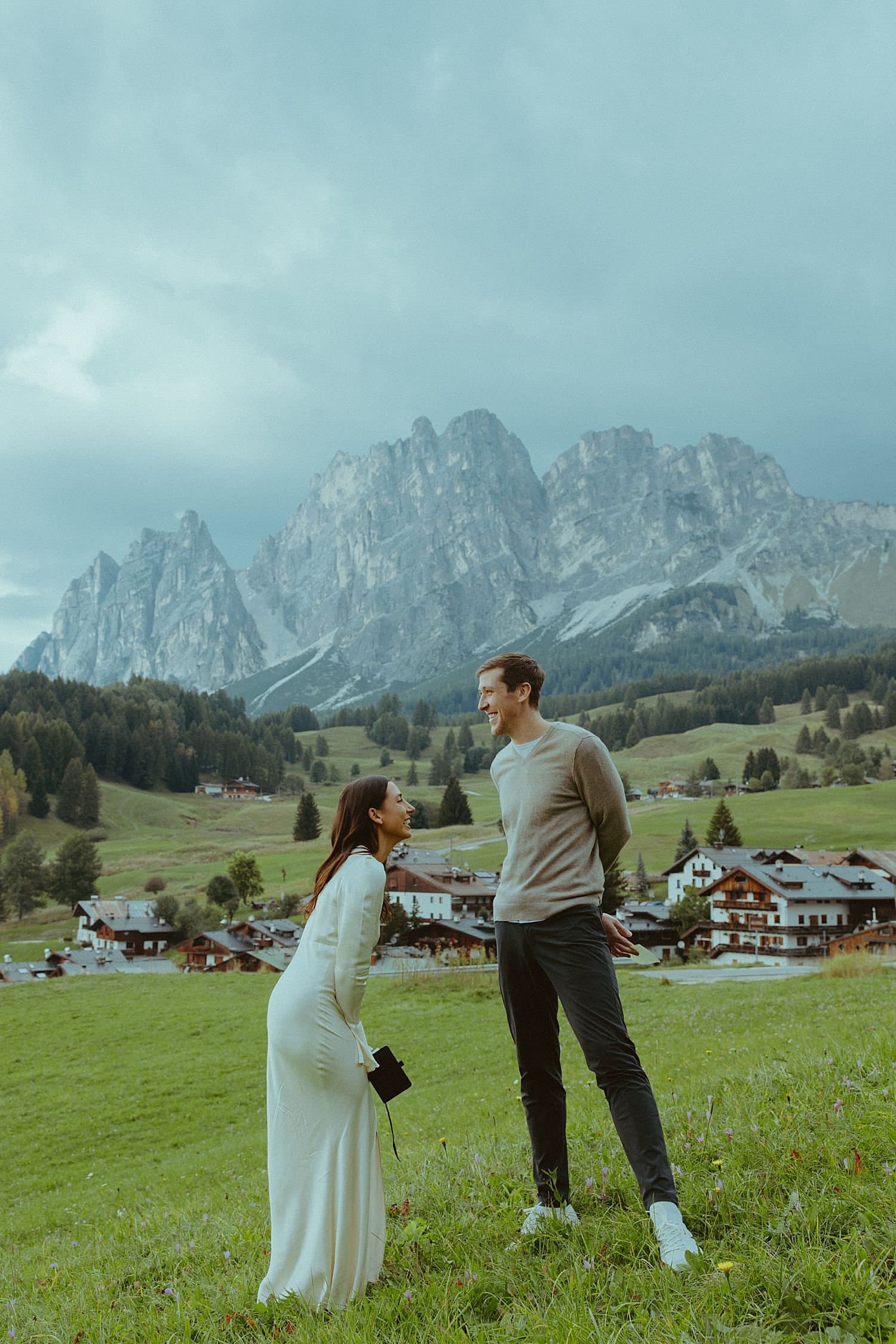 Elopement couple on a green alpine meadow with village chalets and rugged Dolomites peaks behind