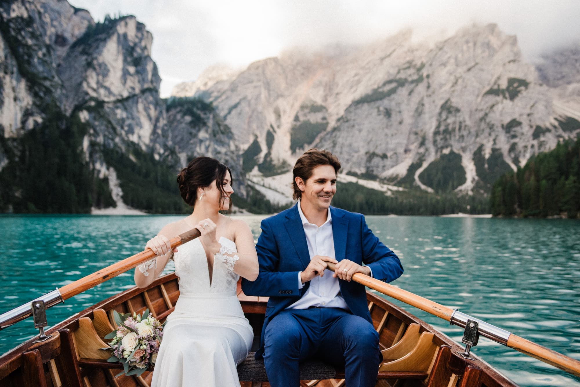 Lago di Braies Wedding & Elopement Photographer: Pricing & Packages Couple rowing a wooden boat across the turquoise water of Lago di Braies beneath the Dolomites