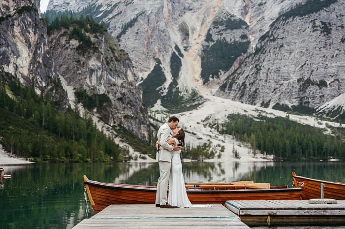 Couple embracing on the wooden dock at Lago di Braies beside a rowing boat, turquoise lake and mountains behind