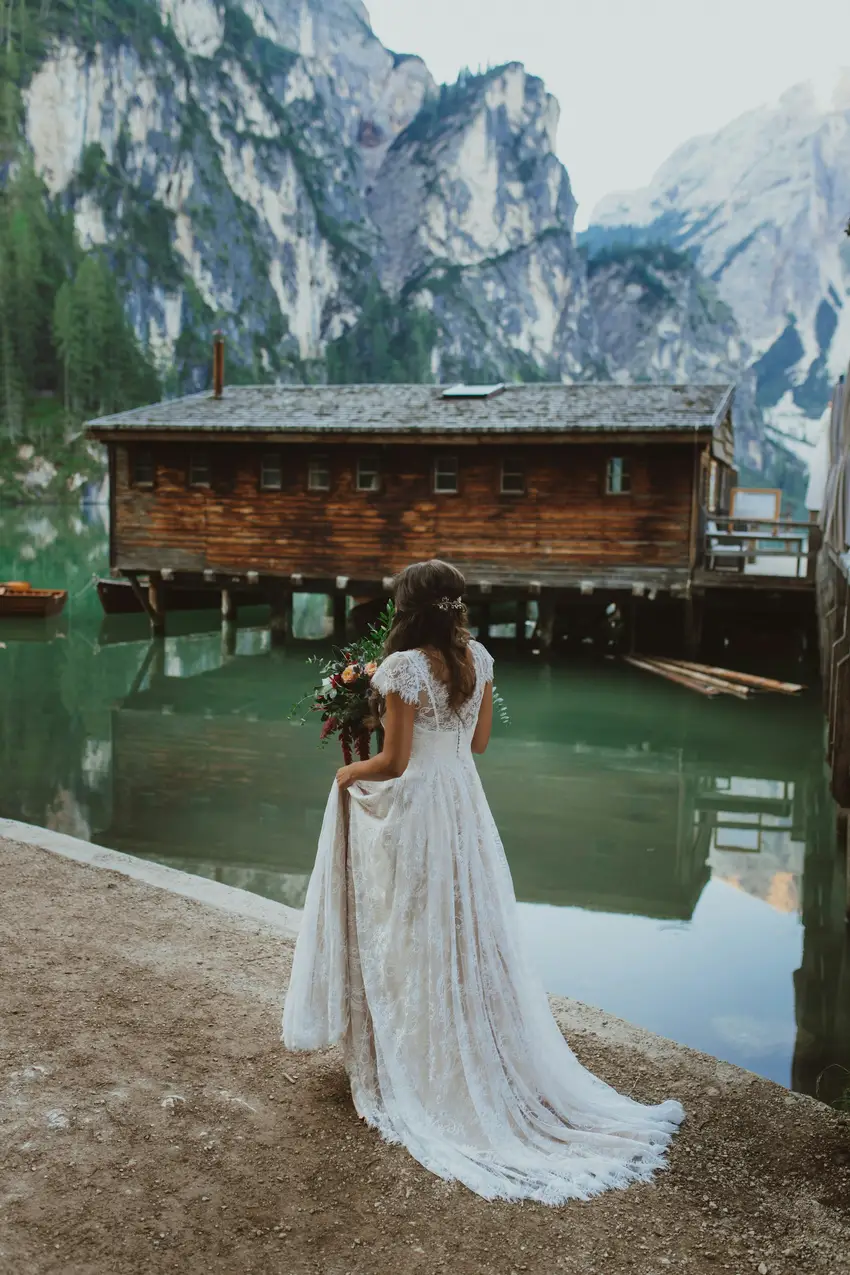 What to Wear to a Mountain Elopement: A Practical Dress Guide Bride in a lace gown standing on the shore of Lago di Braies facing its wooden boathouse
